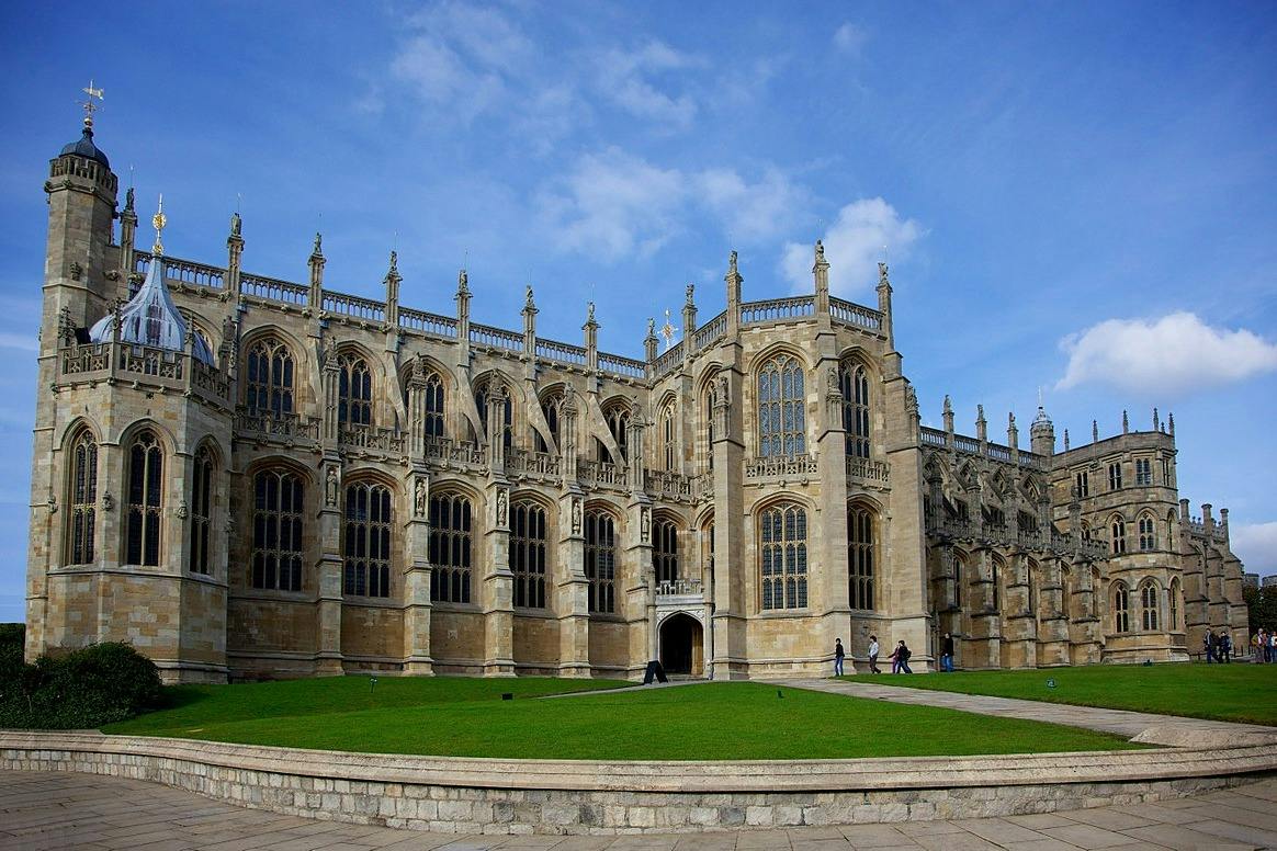 A historic stone building with large Gothic-style windows and spires, surrounded by a green lawn under a blue sky.