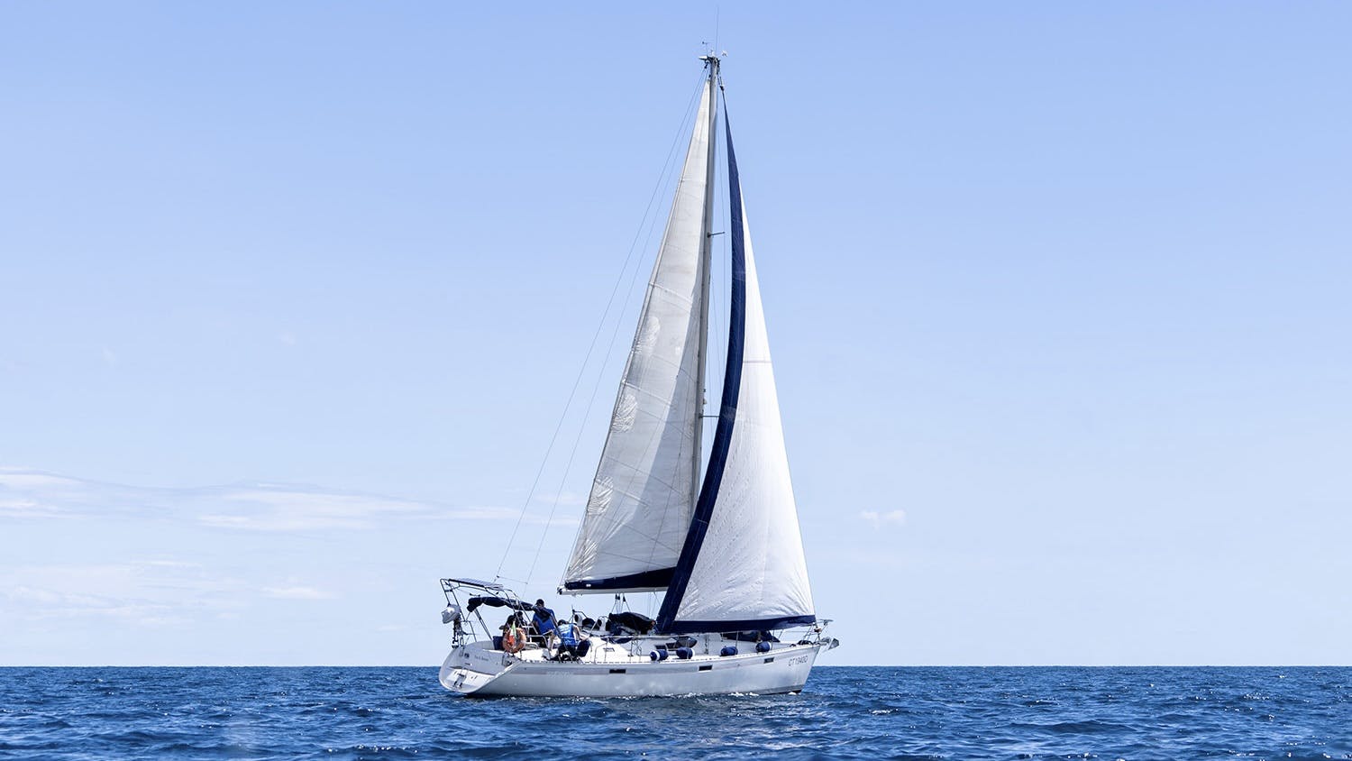 Plachetnice s bílými plachtami se plaví po klidném oceánu pod jasnou modrou oblohou; na palubě lodi jsou vidět lidé.