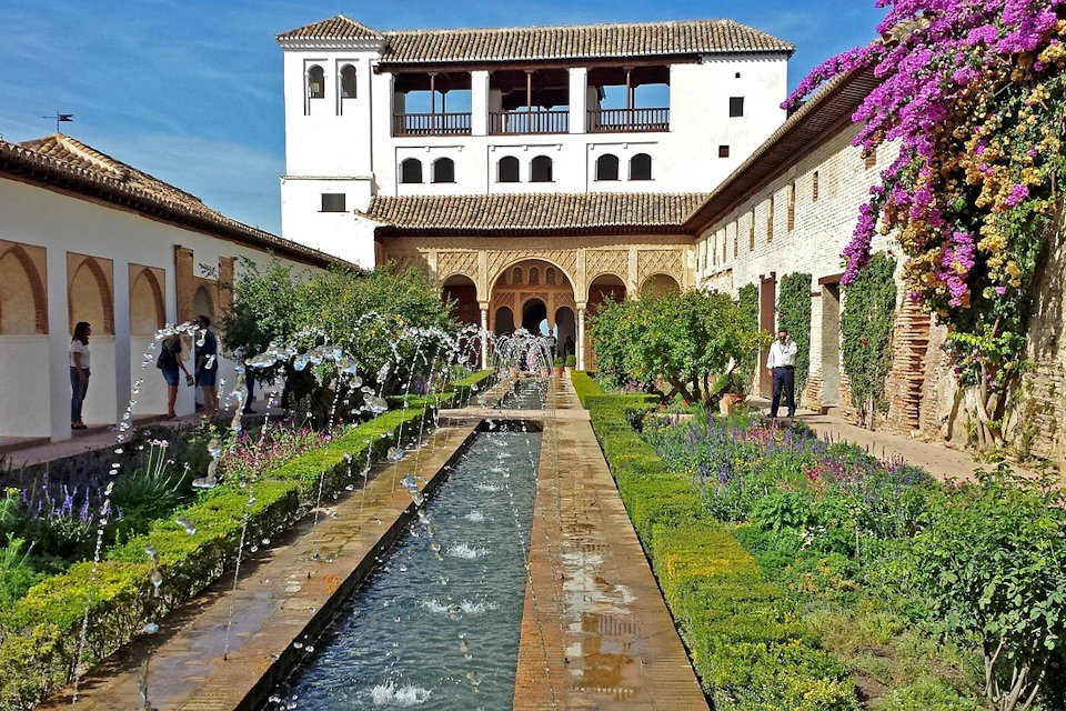 A courtyard with a long fountain, greenery, and a historic building with arches and balconies under a blue sky.