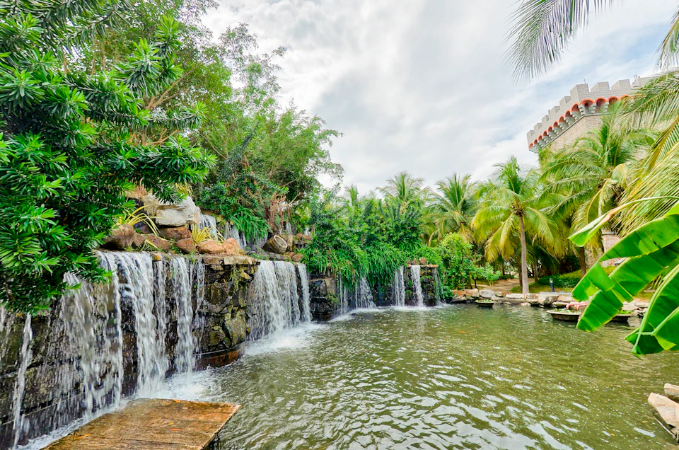A tropical waterfall cascading into a pond surrounded by lush greenery and palm trees under a cloudy sky.
