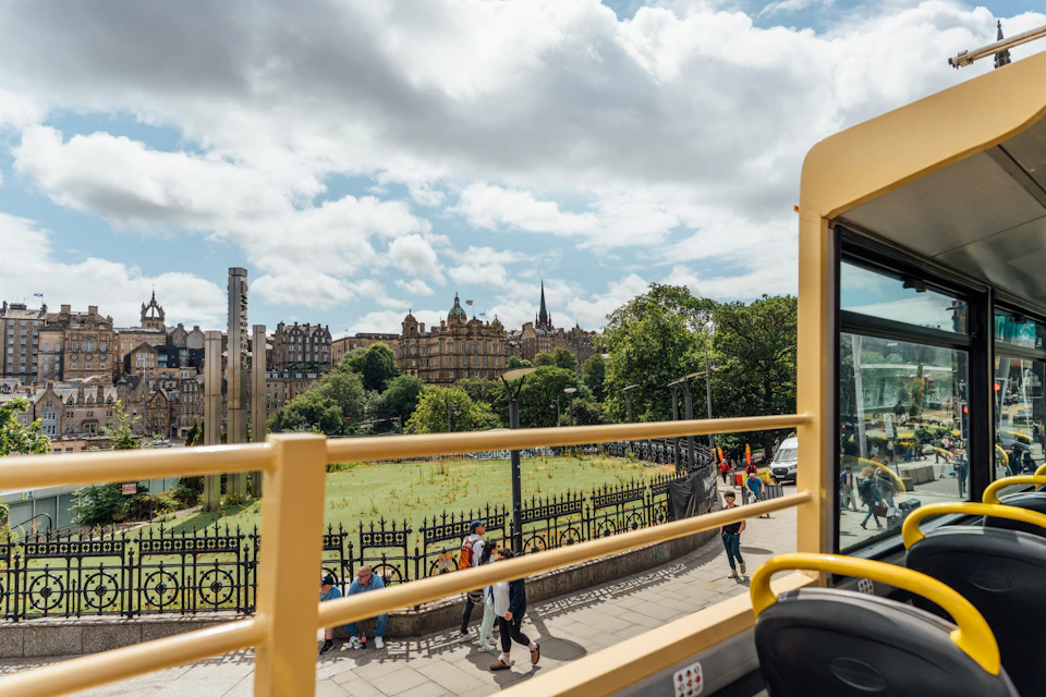 View from an open-top bus overlooking a park, pedestrians, a historic building, and a partly cloudy sky.
