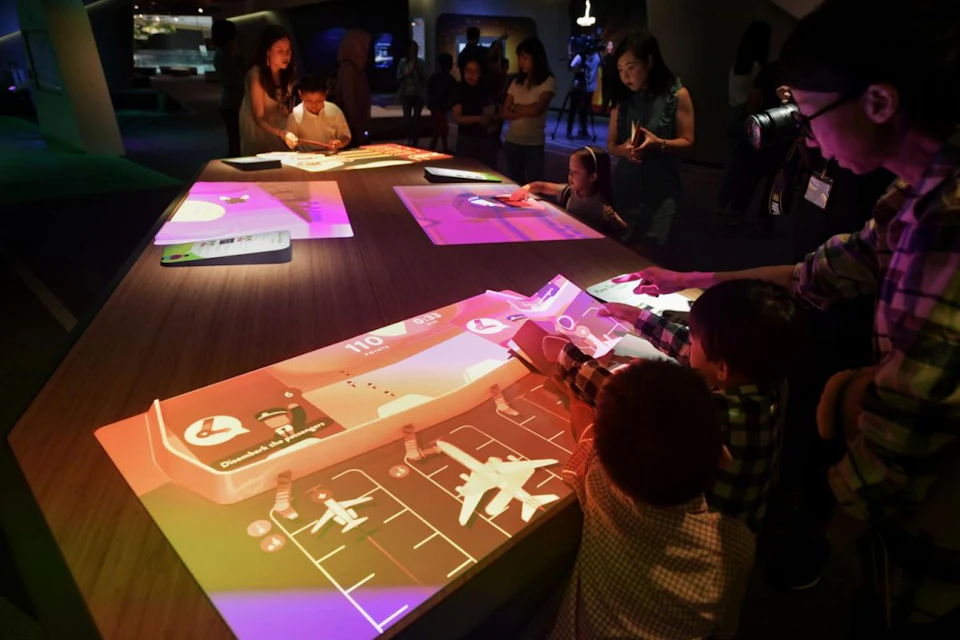 People interacting with illuminated touchscreens displaying graphics and information, in a dimly lit room.