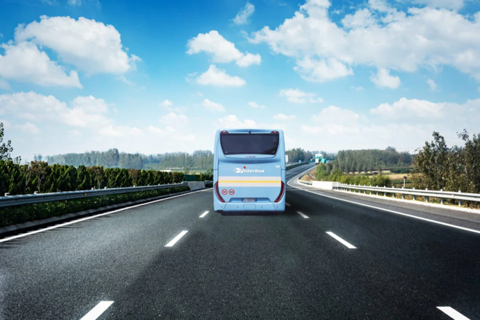 A blue and white bus drives on a highway under a partly cloudy sky, surrounded by trees and fields.