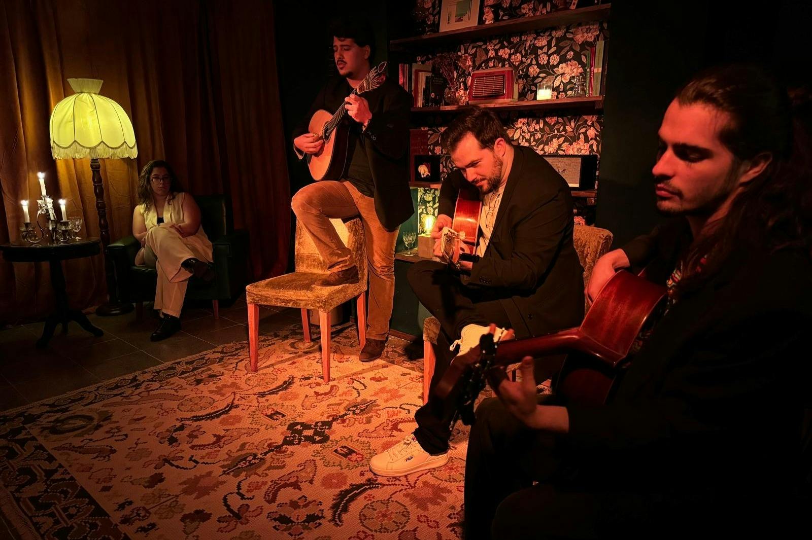 Τρεις άνδρες παίζουν κιθάρες σε ένα αμυδρά φωτισμένο δωμάτιο με ένα χαλί με μοτίβα και ένα ράφι στο παρασκήνιο.