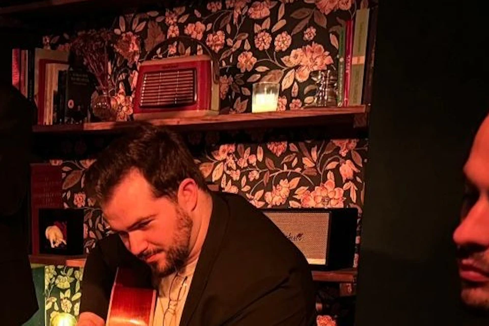 Three men playing guitars in a dimly lit room with a patterned rug and a bookshelf in the background.