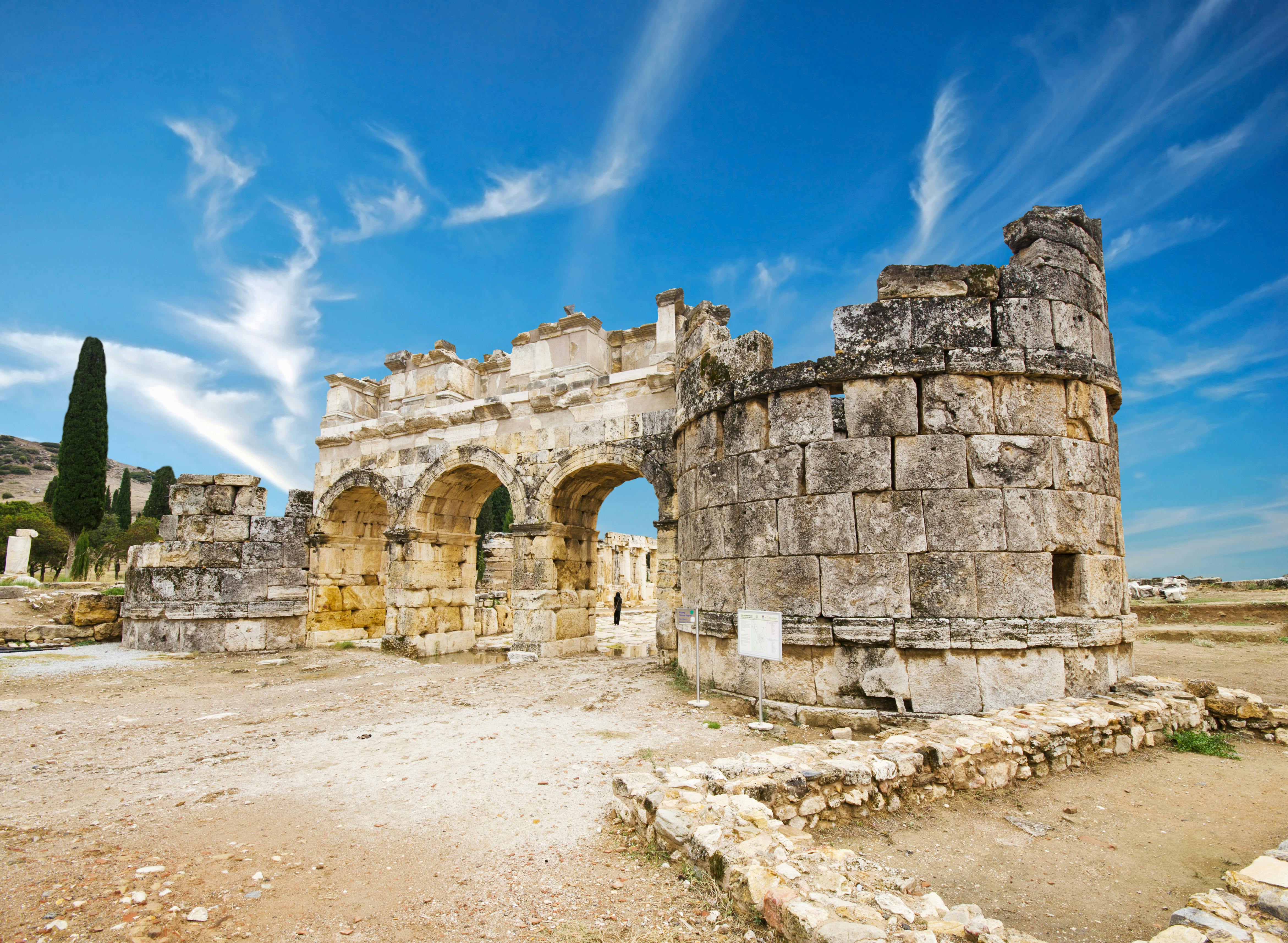 Ancient stone ruins with arches under a bright blue sky with wispy clouds. The ground is rocky and worn.
