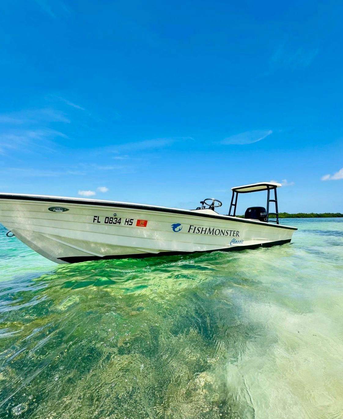 Un bateau à moteur blanc portant l'inscription "FishMonster" flotte dans une eau turquoise claire et peu profonde, sous un ciel d'un bleu éclatant.