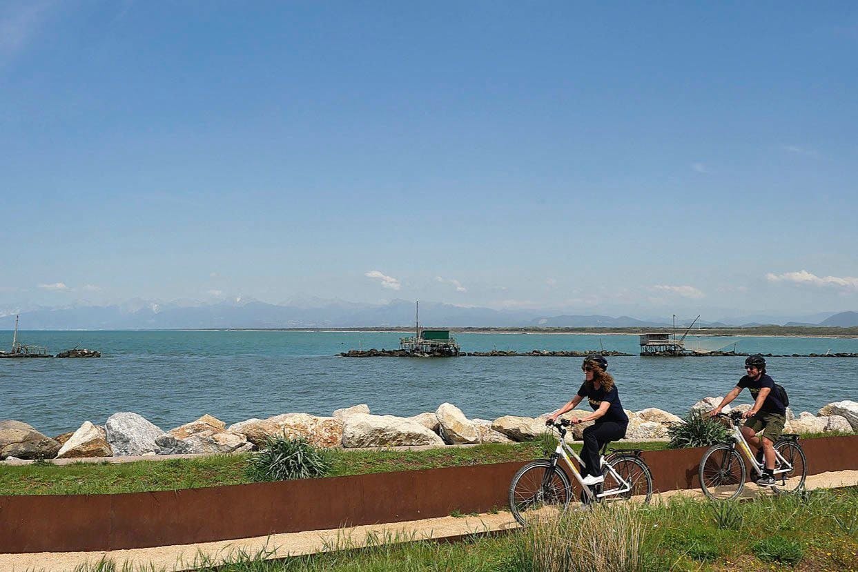 A woman rides a bicycle along a coastal path with large rocks and calm sea water, under a clear blue sky.
