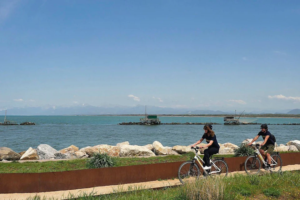 A woman rides a bicycle along a coastal path with large rocks and calm sea water, under a clear blue sky.
