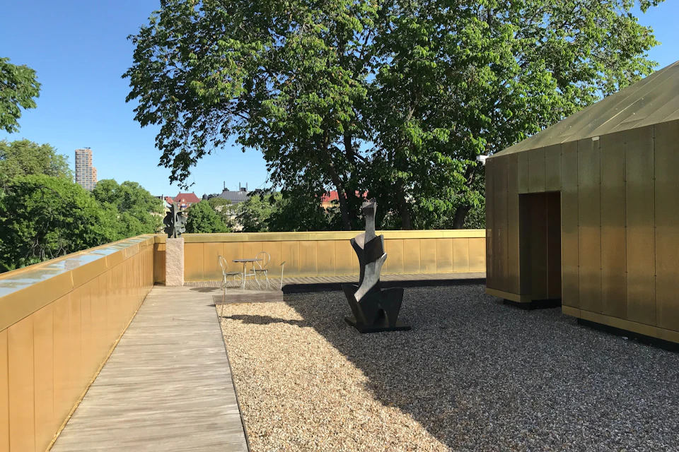A rooftop with a modern sculpture, gravel, and a small table set, surrounded by trees and golden walls, under a clear sky.