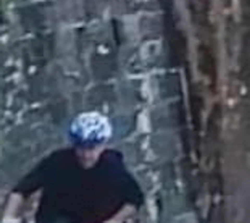 Three people with helmets riding bicycles on a cobblestone path beside a moss-covered wall with trees in the background.