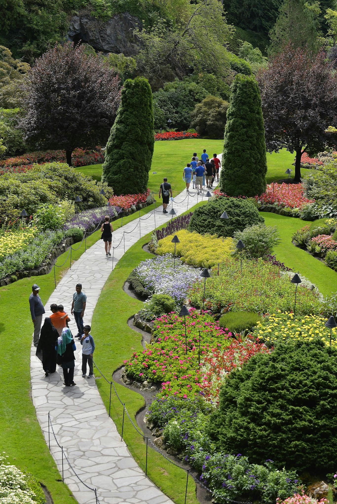 Οι άνθρωποι περπατούν σε πέτρινα μονοπάτια σε έναν ζωντανό κήπο με περιποιημένους χλοοτάπητες, πολύχρωμα παρτέρια και ψηλά δέντρα.