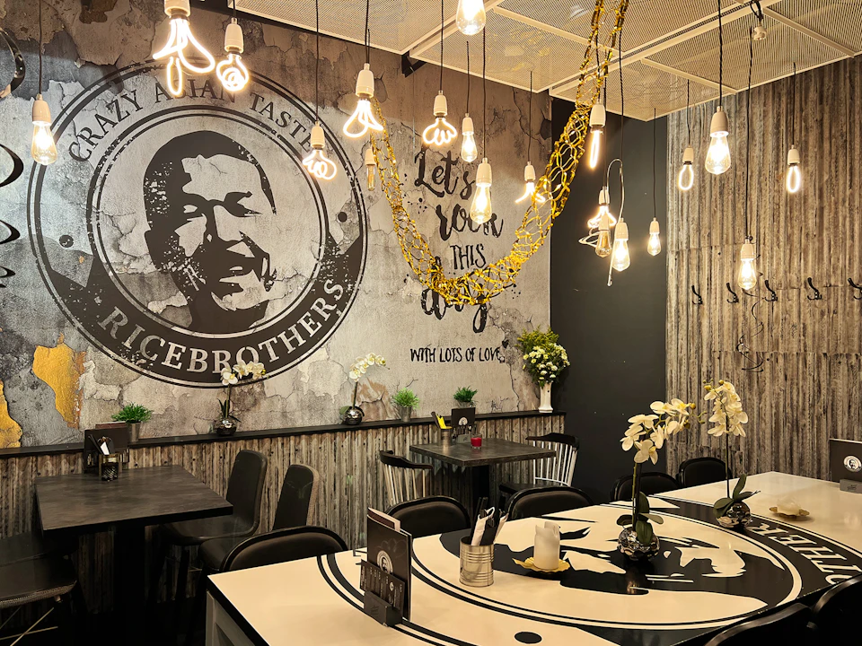 A restaurant interior with hanging pendant lights, rustic decor, wall art, and floral arrangements on tables and shelves.
