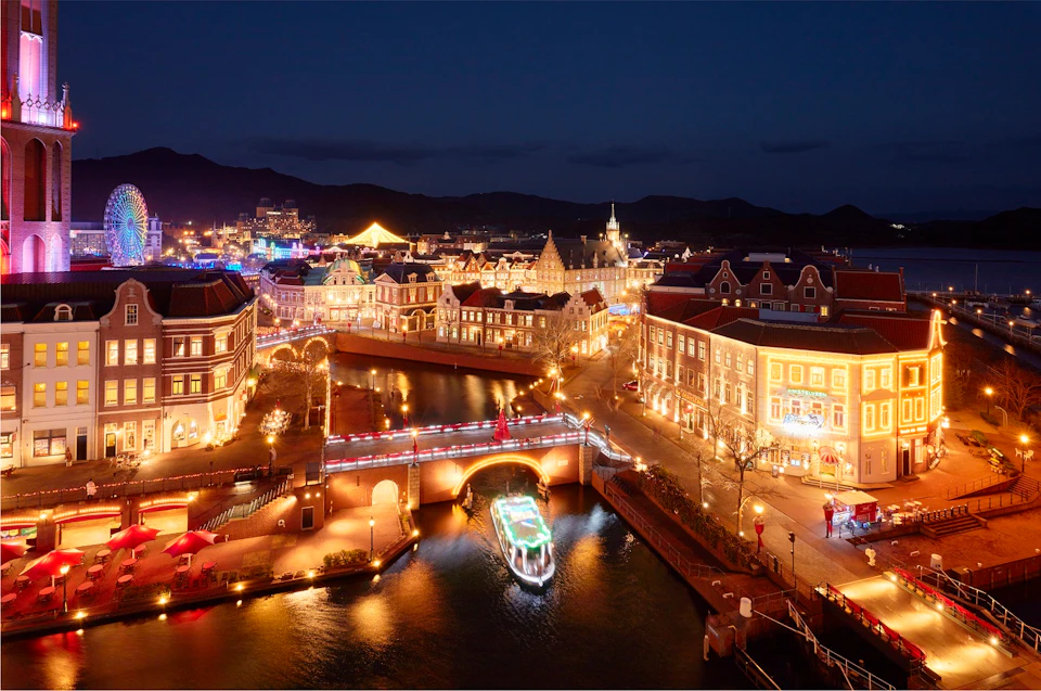 A nighttime cityscape with illuminated buildings, a lit canal, and a boat passing under a bridge.