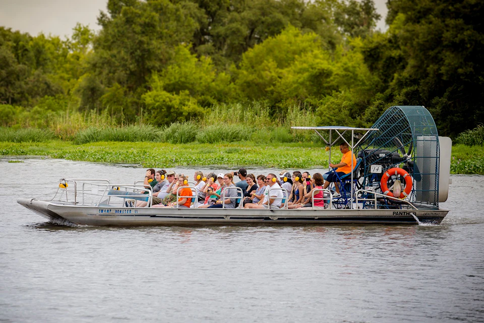 A group of people seated on an airboat cruising through a river, surrounded by greenery and trees.