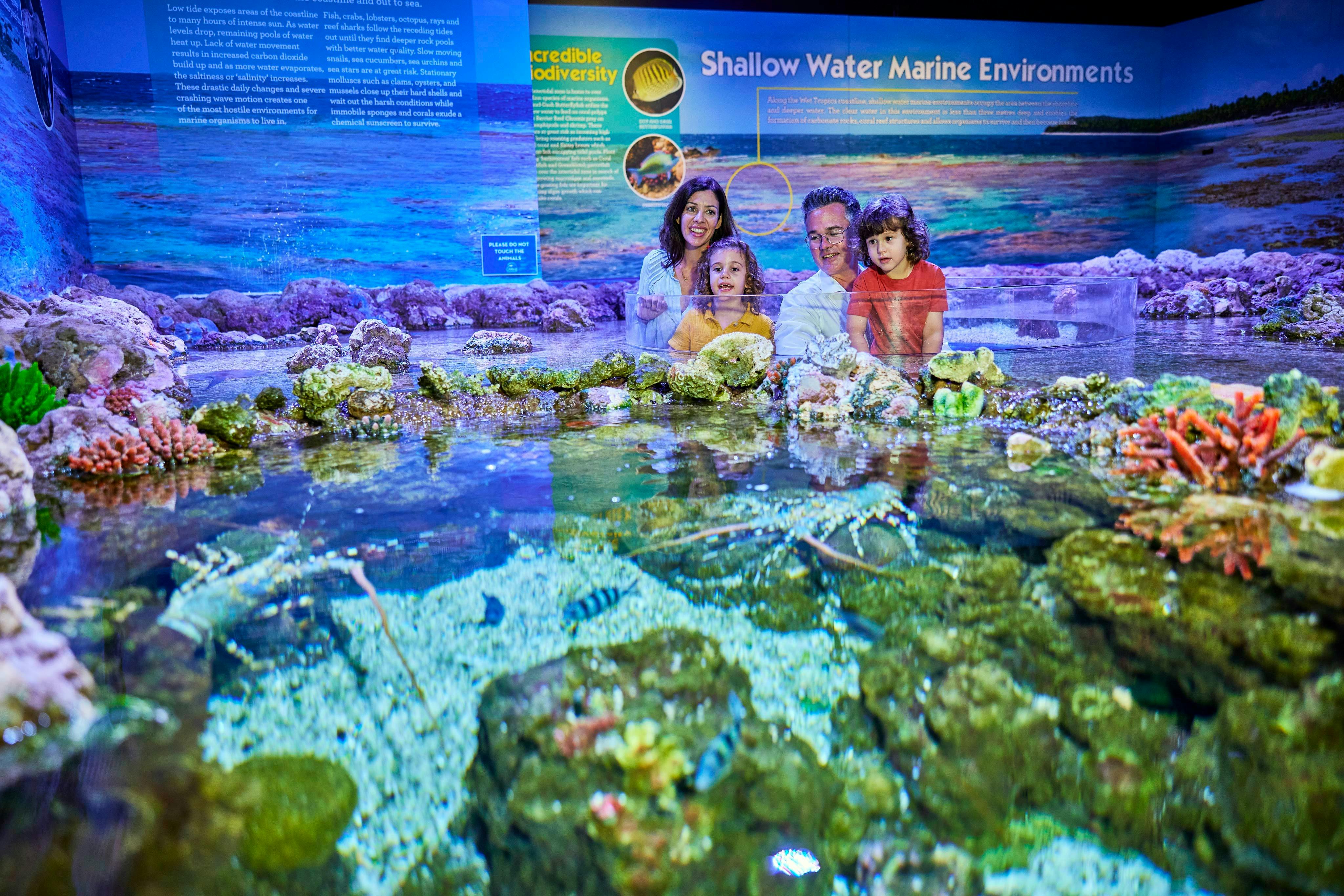 Czteroosobowa rodzina pozuje za płytkowodnym eksponatem morskim z koralami, skałami i rybami w sali wystawowej akwarium.