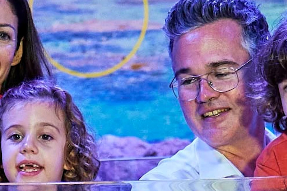 A family of four poses behind a shallow water marine exhibit with coral, rocks, and fish in an aquarium display room.