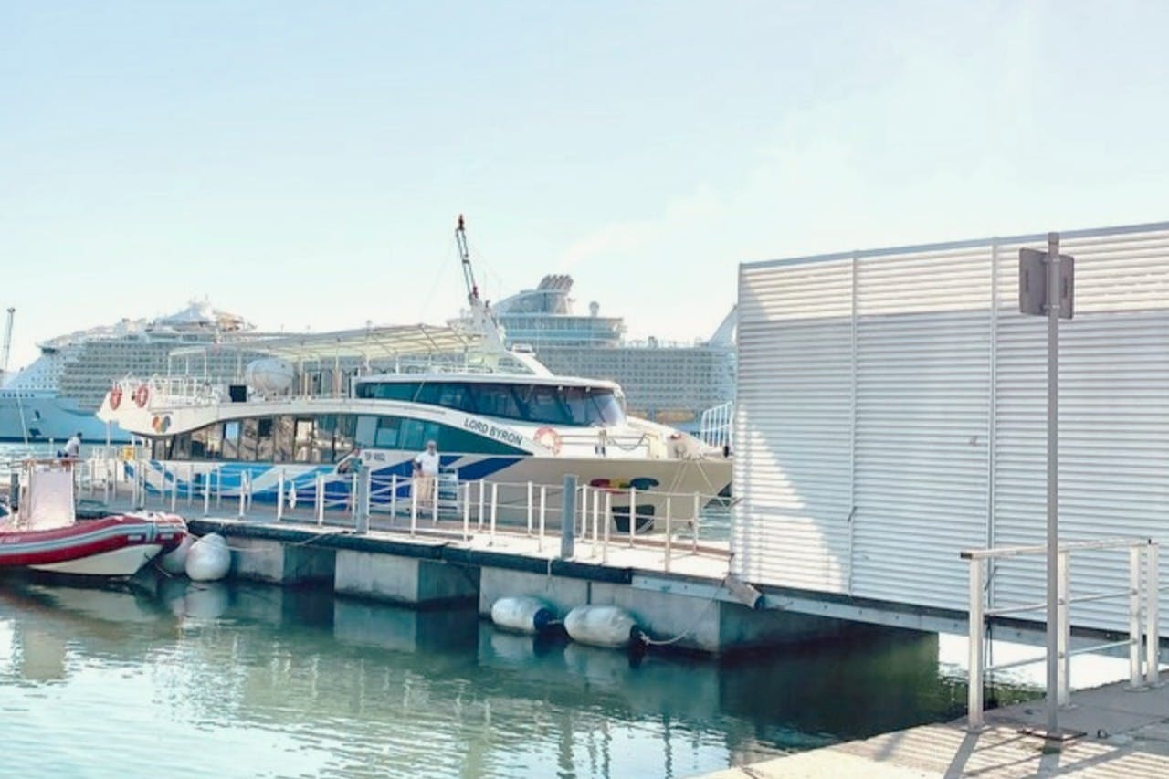 Ένα ελλιμενισμένο επιβατηγό πλοίο με το όνομα «Λόρδος Βύρων» κοντά σε μια προβλήτα, με ένα μεγάλο κρουαζιερόπλοιο ορατό στο παρασκήνιο.