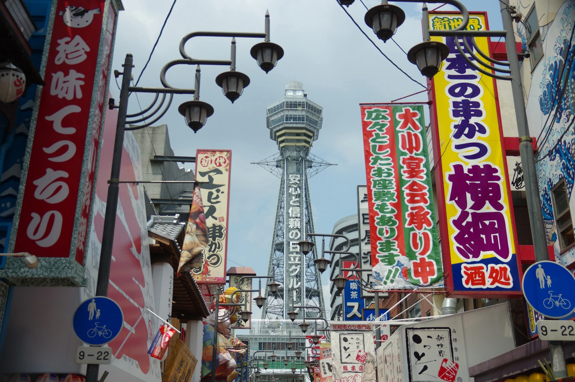 A bustling street with colorful signs and banners, featuring a tall tower in the background, under a cloudy sky.