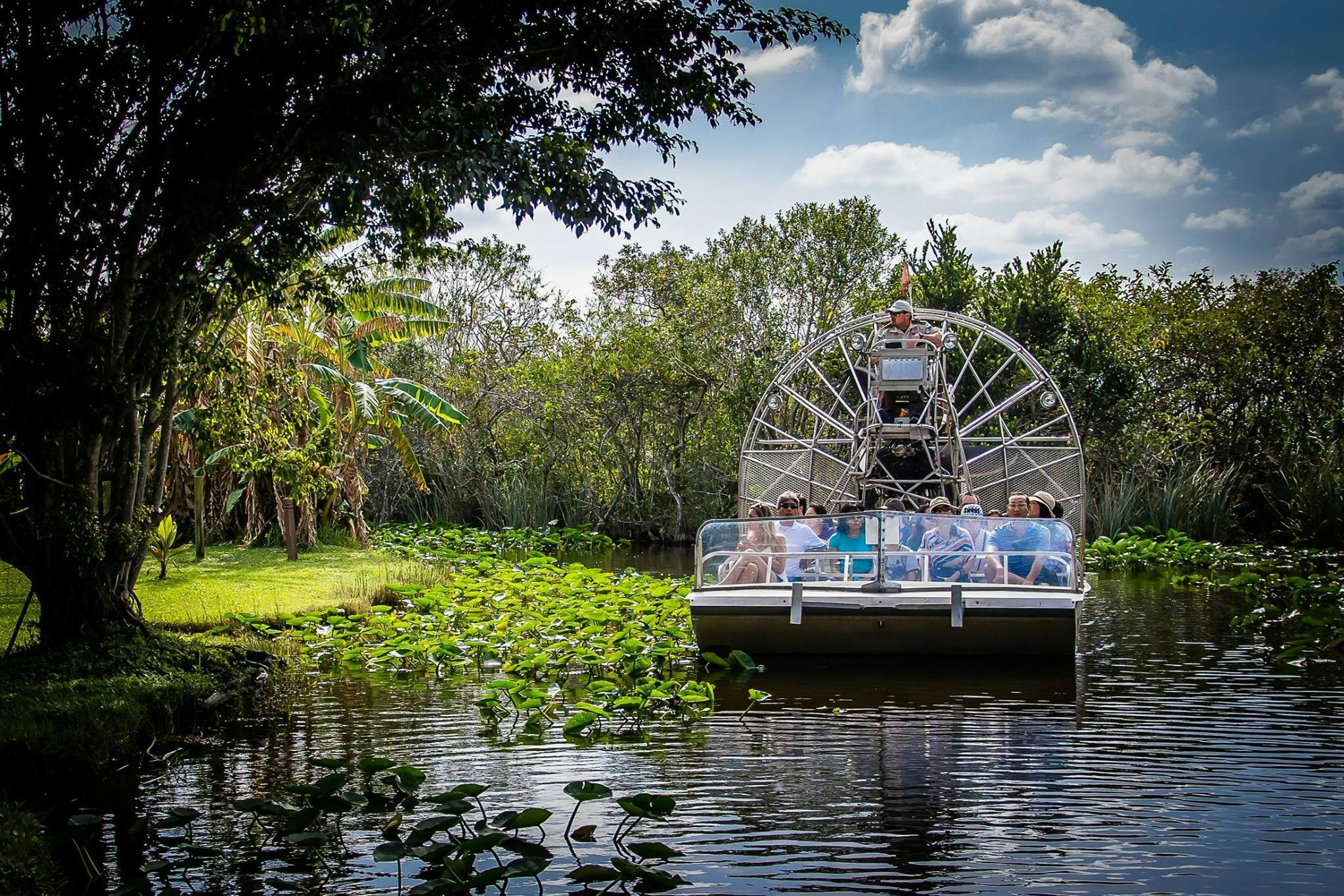 An airboat with passengers navigates a waterway surrounded by lush vegetation under a partly cloudy sky.
