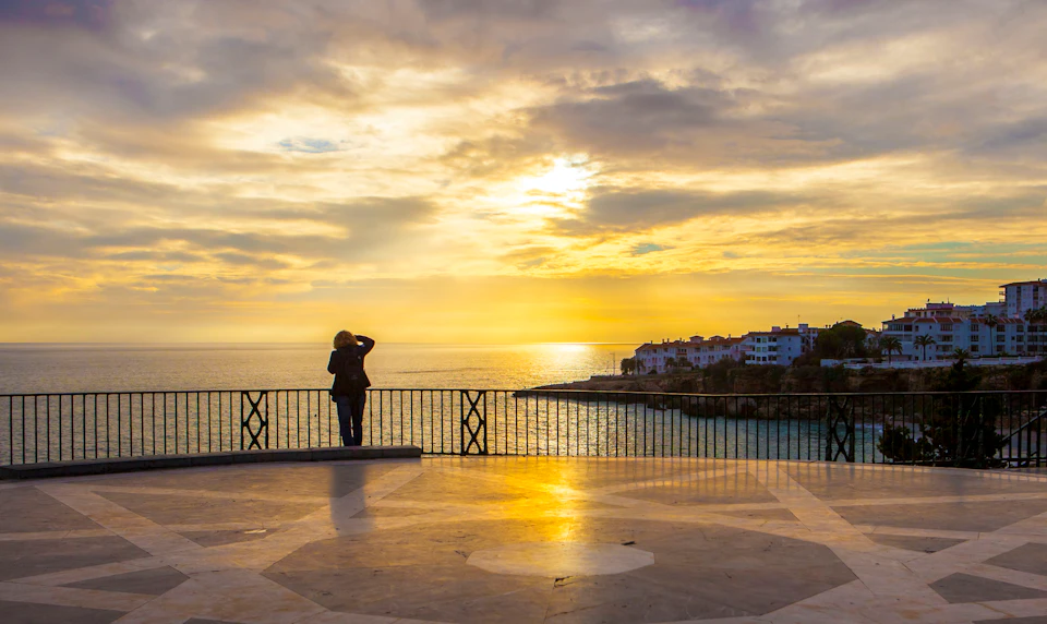 A person stands at a railing, looking out over the ocean at sunset. Buildings are visible on the coastline in the background.