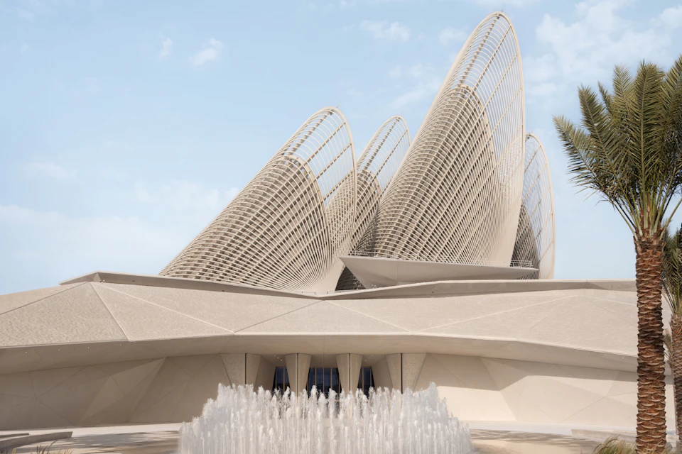 A modern architectural building with curved, lattice-like structures and a fountain in the foreground under a clear sky.