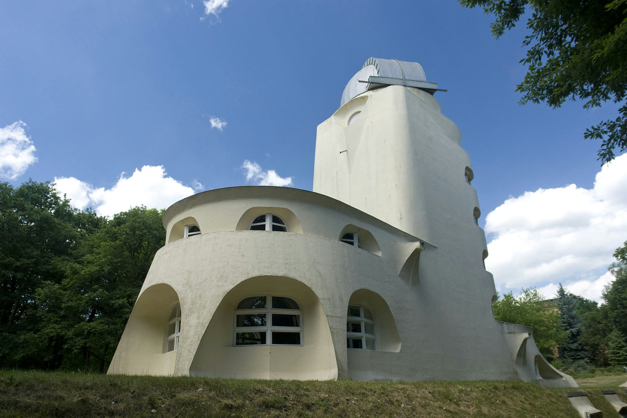 Ein weißes, modernistisches Gebäude mit gewölbten Fenstern und einem zylindrischen Turm vor einer Kulisse aus Bäumen und blauem Himmel.