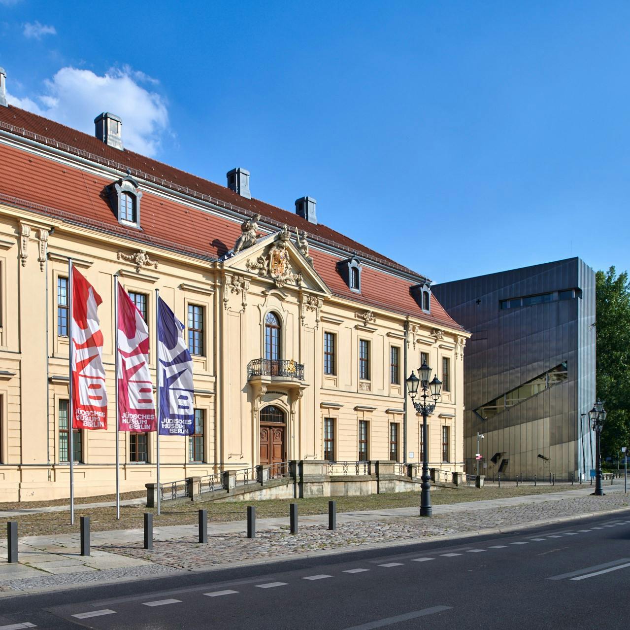 Historická budova s červenou střechou a klenutými okny vedle moderní stavby. Venku visí tři vlajky. Jasná modrá obloha.