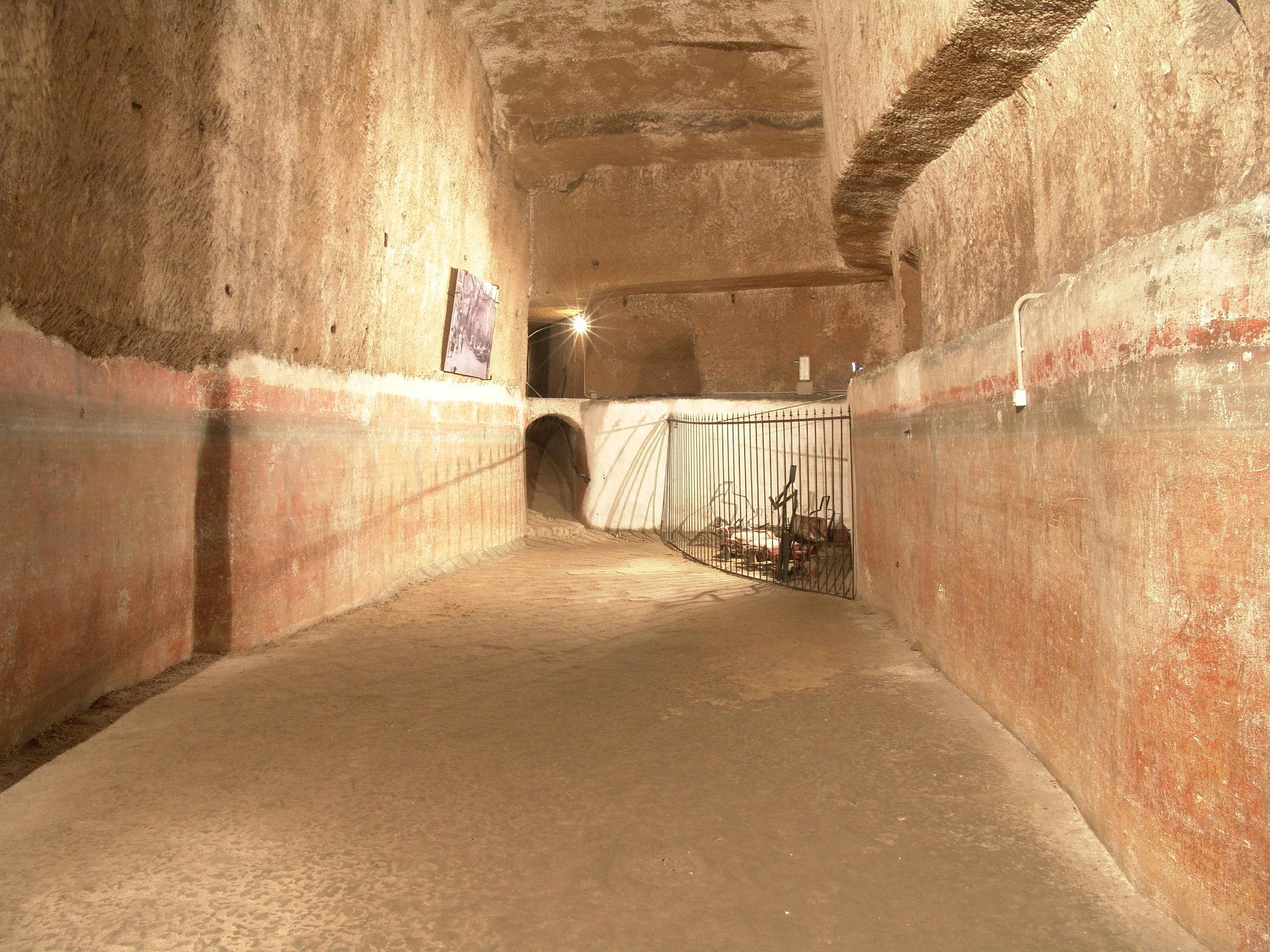 Un couloir souterrain faiblement éclairé avec des murs en pierre, des bandes horizontales rouges et brunes, une arche, une porte et des images encadrées sur le mur.