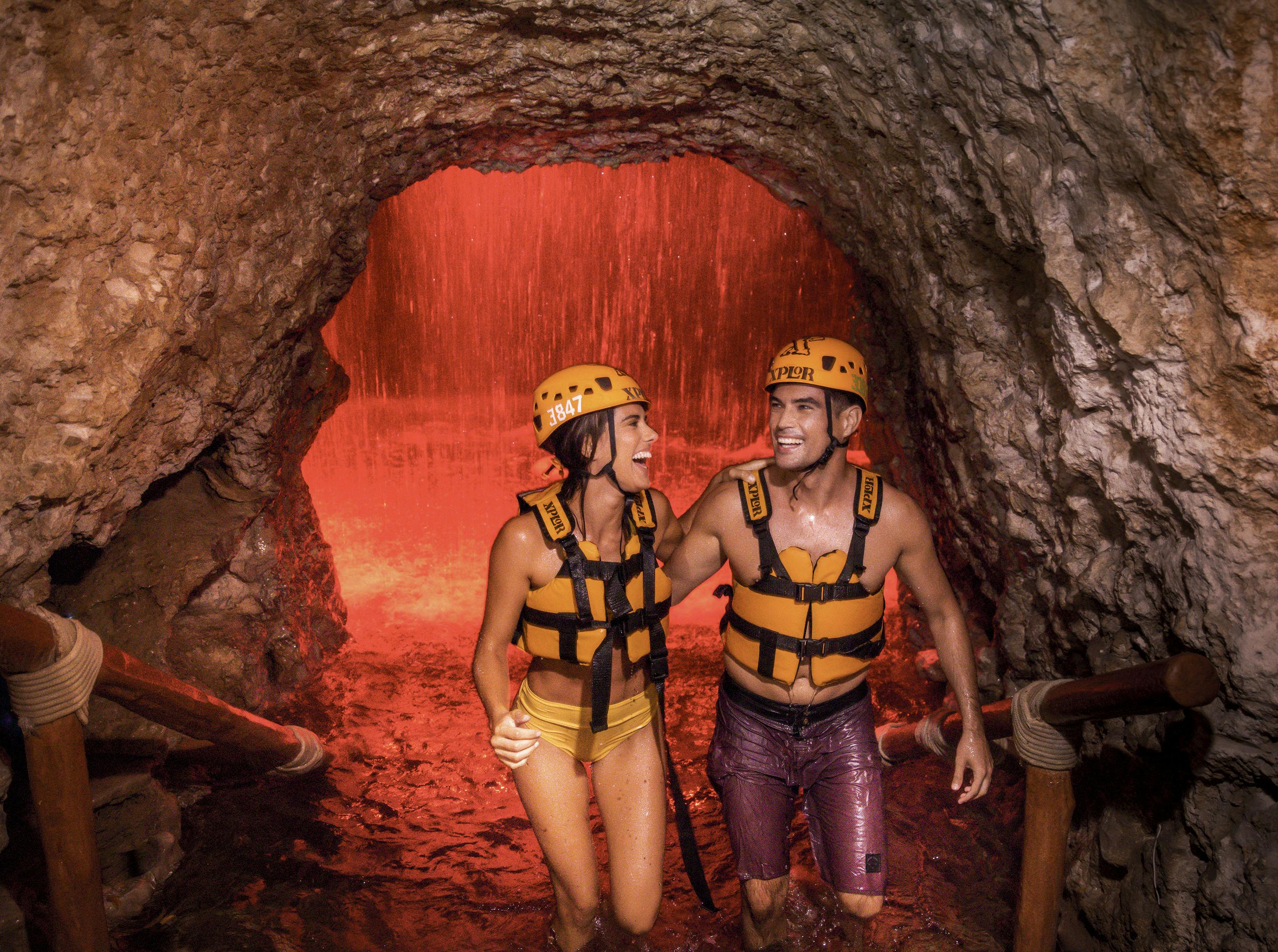 Un hombre y una mujer sonrientes con casco y chaleco salvavidas caminan por una cueva rocosa con una cascada roja al fondo.