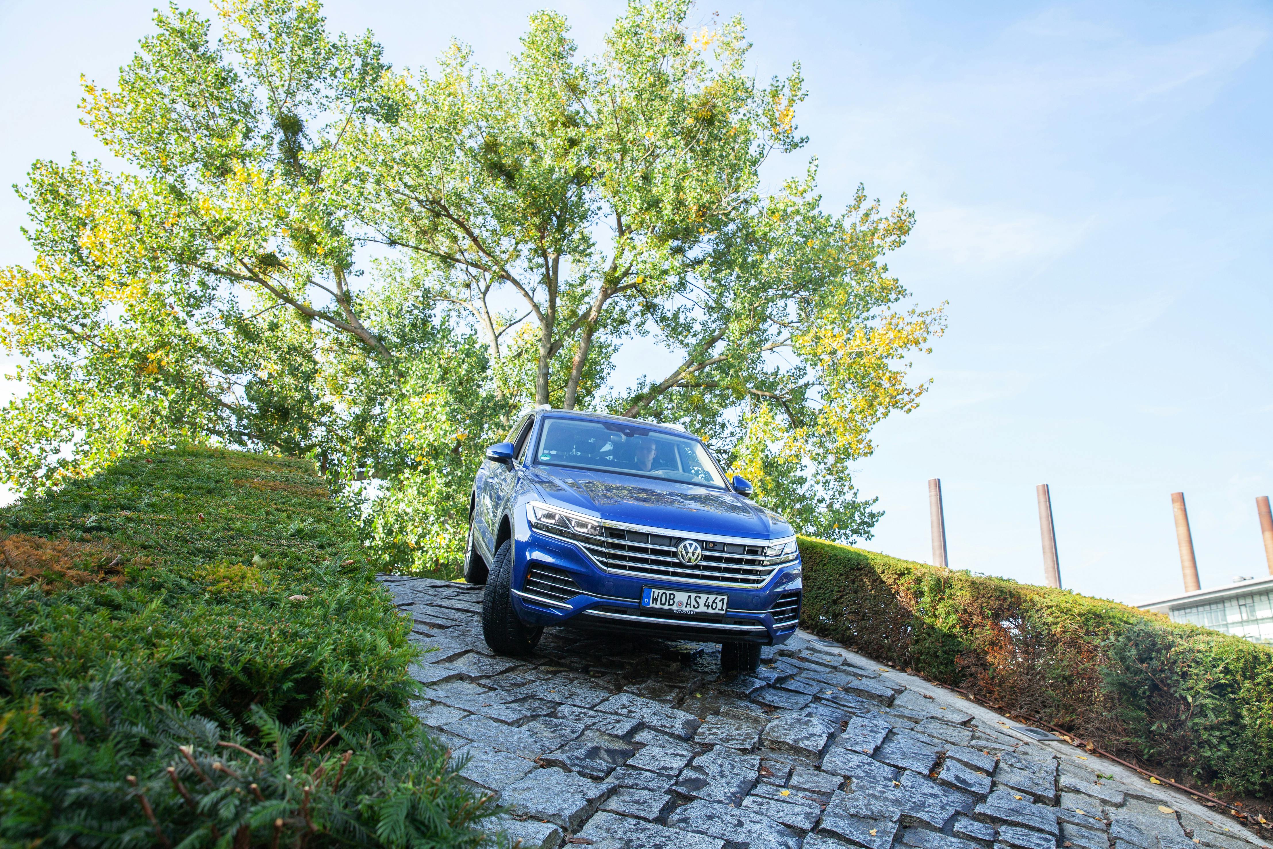 A blue SUV navigates a steep, cobblestone path with green bushes and tall trees in the background.