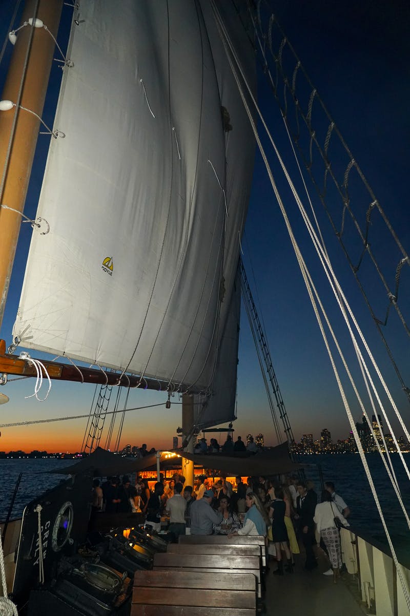 Ένα ιστιοφόρο με μεγάλα πανιά είναι αγκυροβολημένο ή πλέει το βράδυ, με τους ανθρώπους στο πλοίο και έναν ορίζοντα της πόλης ορατό στο παρασκήνιο.