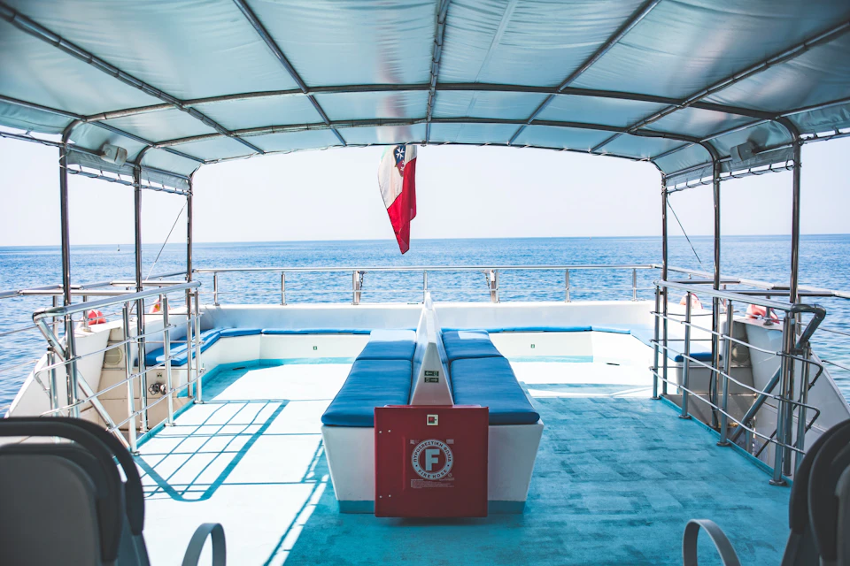 A covered deck of a boat with seating, a flag at the rear, and an ocean view under a clear sky.