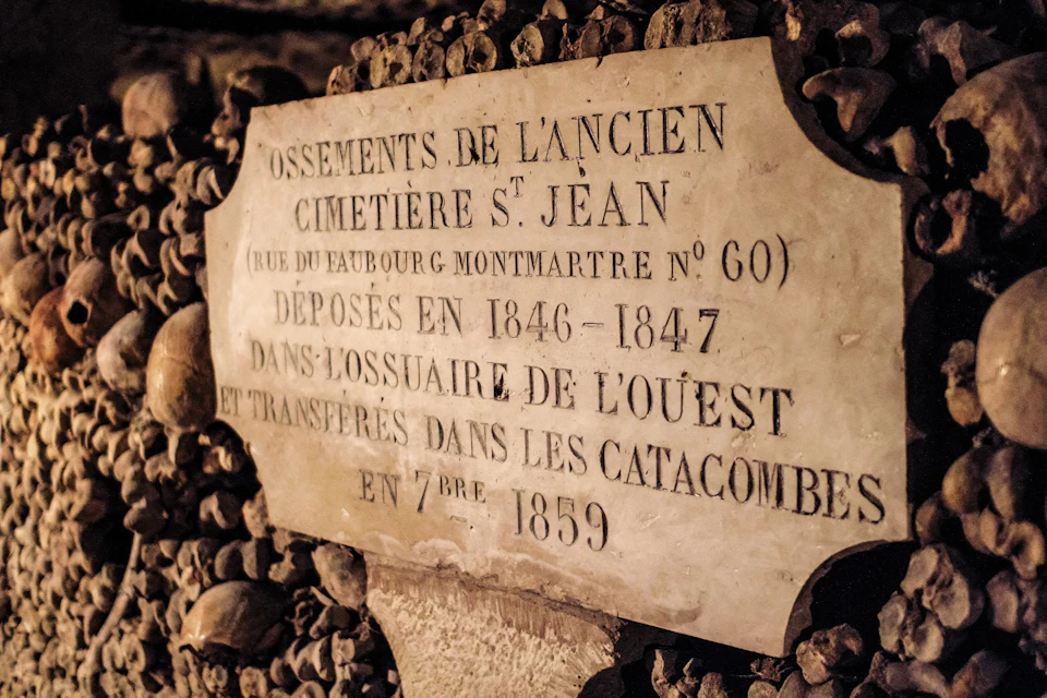 A plaque in the Catacombs of Paris, surrounded by stacked bones, with French text detailing historical burial information.