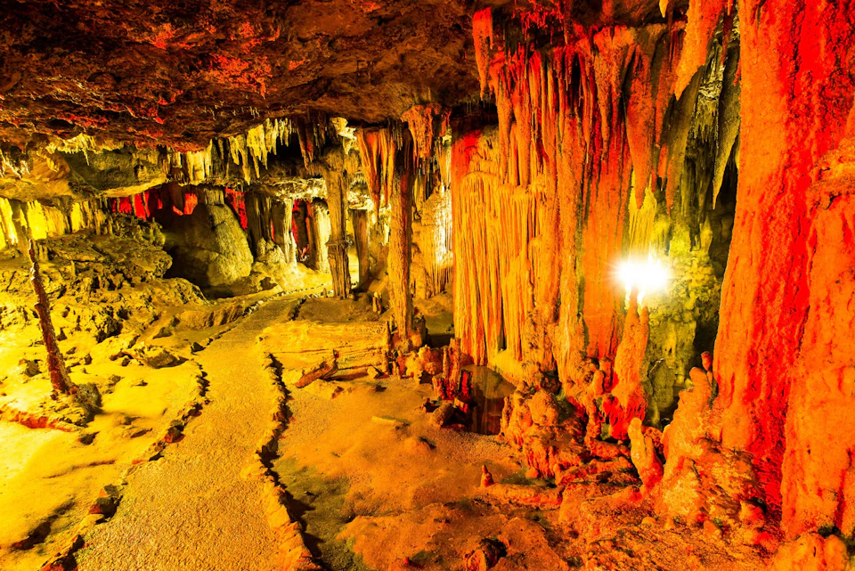 A cave interior featuring numerous stalactites and stalagmites illuminated by warm golden and orange lights.