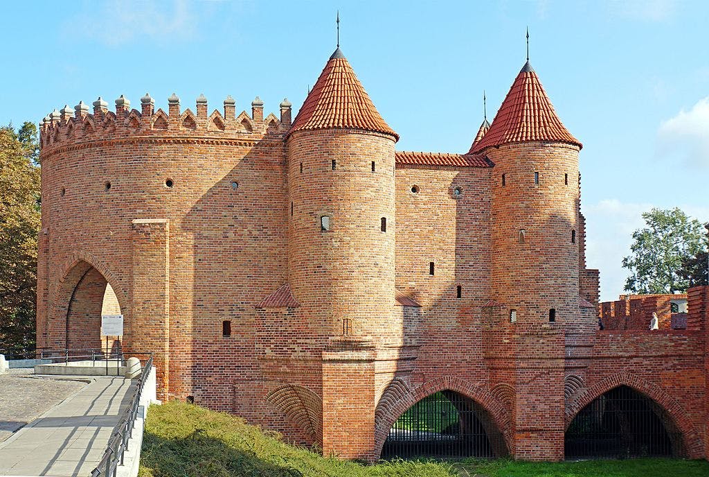 Historická cihlová pevnost s klenutými branami, několika věžemi a cimbuřím pod modrou oblohou.