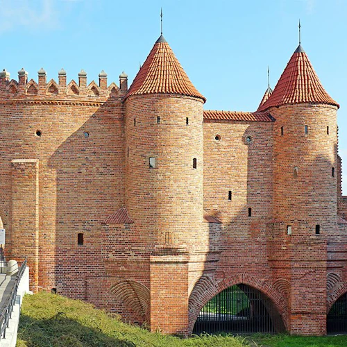En historisk fästning i tegel med välvda portar, flera torn och krenellerade murar under en blå himmel.