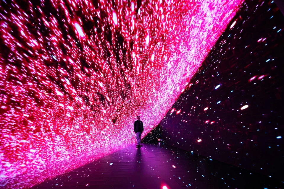 A person walks through a tunnel illuminated by vivid pink and red lights, resembling a starry or cosmic scene.