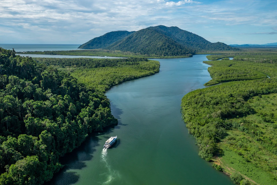 A boat cruises along a river surrounded by dense greenery, with forested hills and a partly cloudy sky in the background.