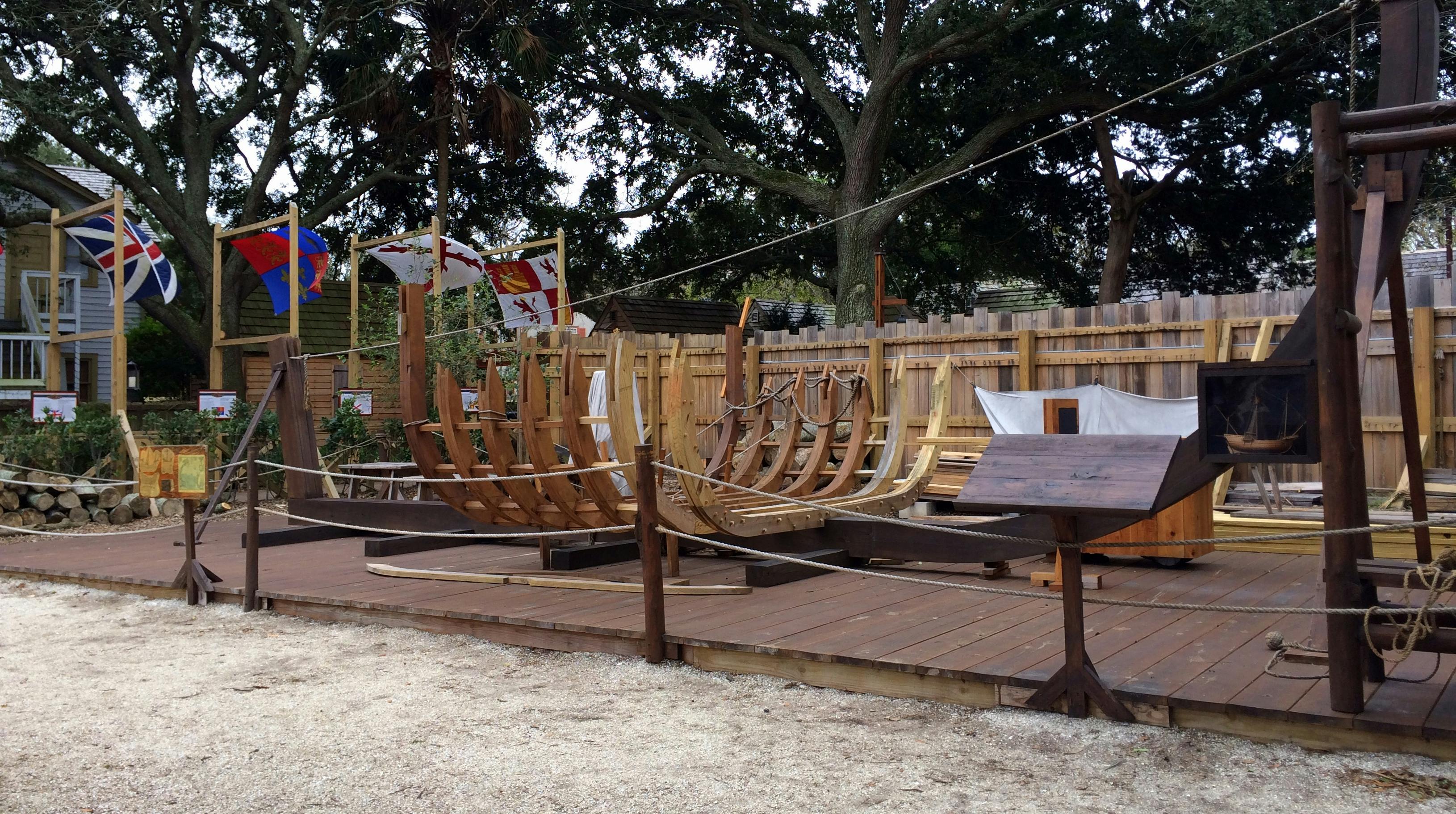 木制甲板上正在建造的木制船舶框架，背景是旗帜和树木。