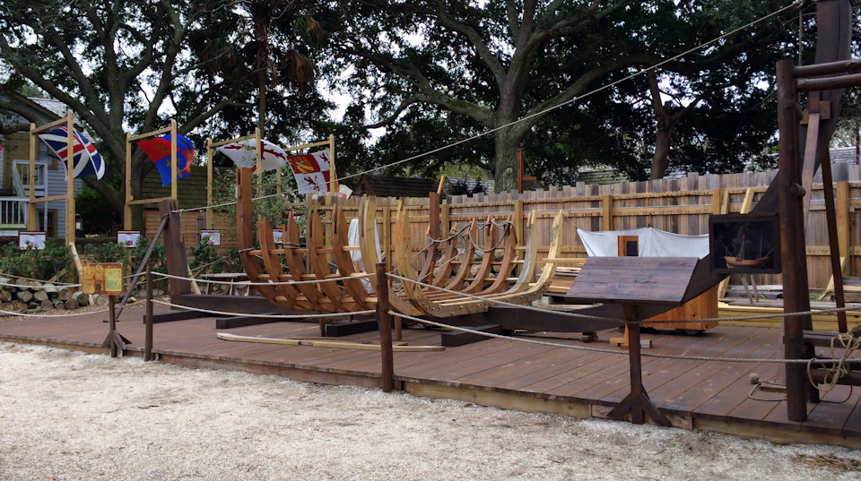 Wooden ship frame under construction on a wooden deck, with flags and trees in the background.