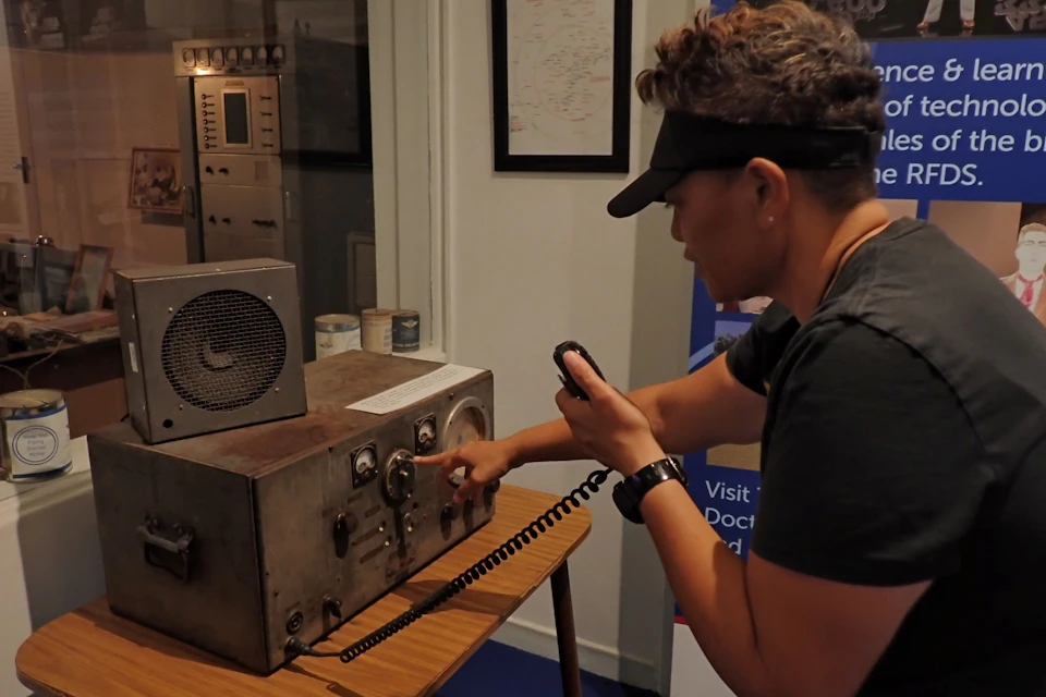 A person operating a vintage communication device with a microphone in hand, standing in a room with historical equipment.