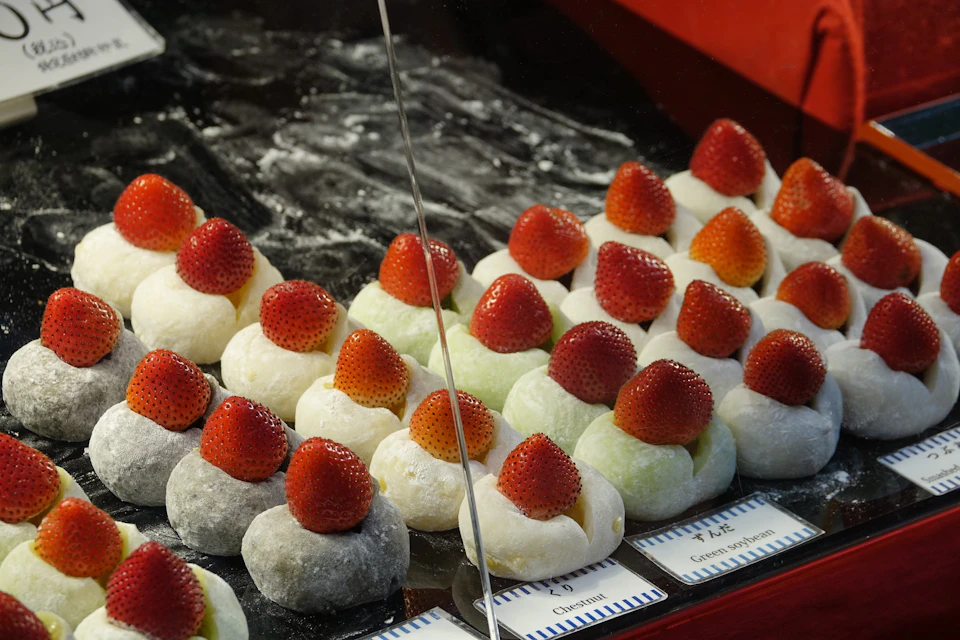 Rows of mochi balls topped with whole strawberries in a display case. The mochi comes in various colors, including white and green.