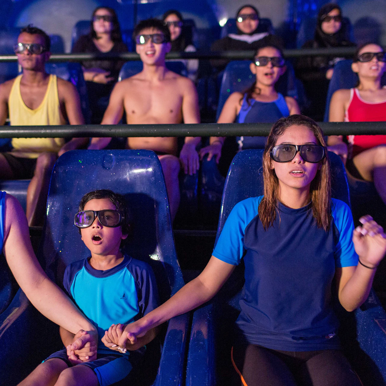 People wearing 3D glasses, seated in a theater-style ride, some holding hands while experiencing an immersive show.