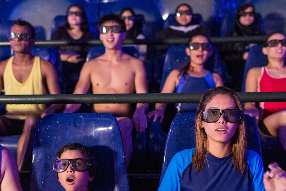 People wearing 3D glasses, seated in a theater-style ride, some holding hands while experiencing an immersive show.