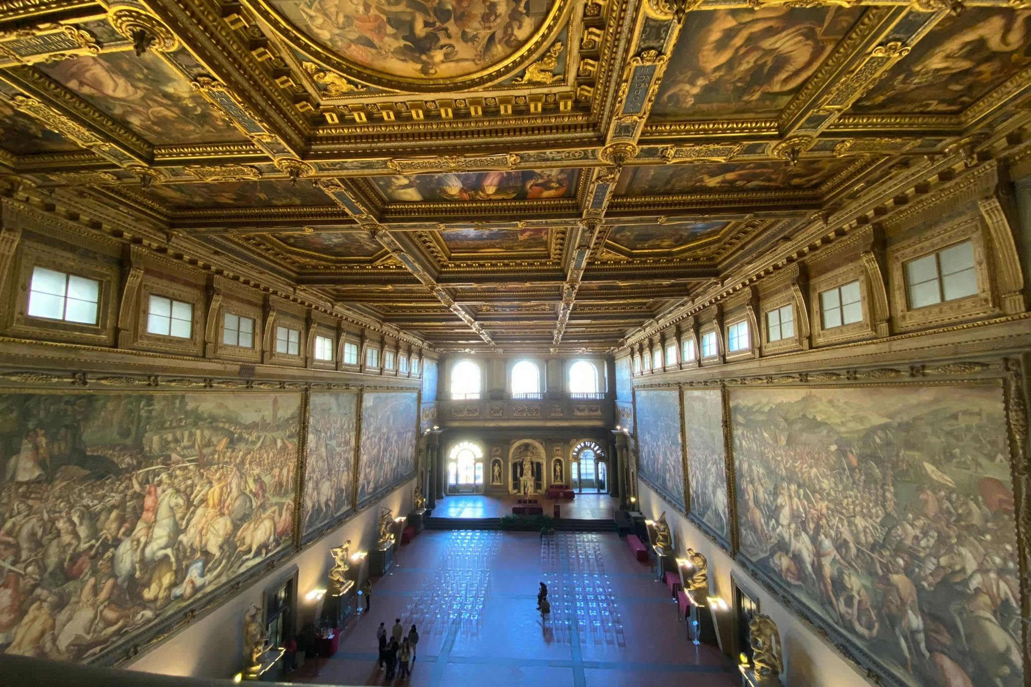 Una gran sala con techos ornamentados y pintados y grandes murales en las paredes. Se ven pocas personas paseando y admirando el espacio.