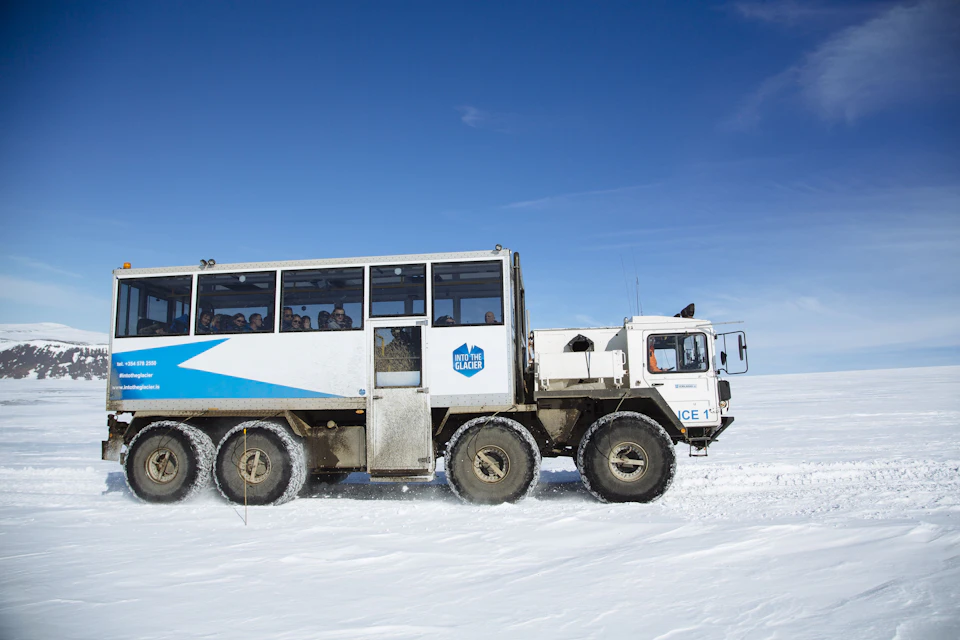 A large, white, off-road vehicle with a blue stripe and "Into the Glacier" logo drives over a snowy terrain under a clear blue sky.
