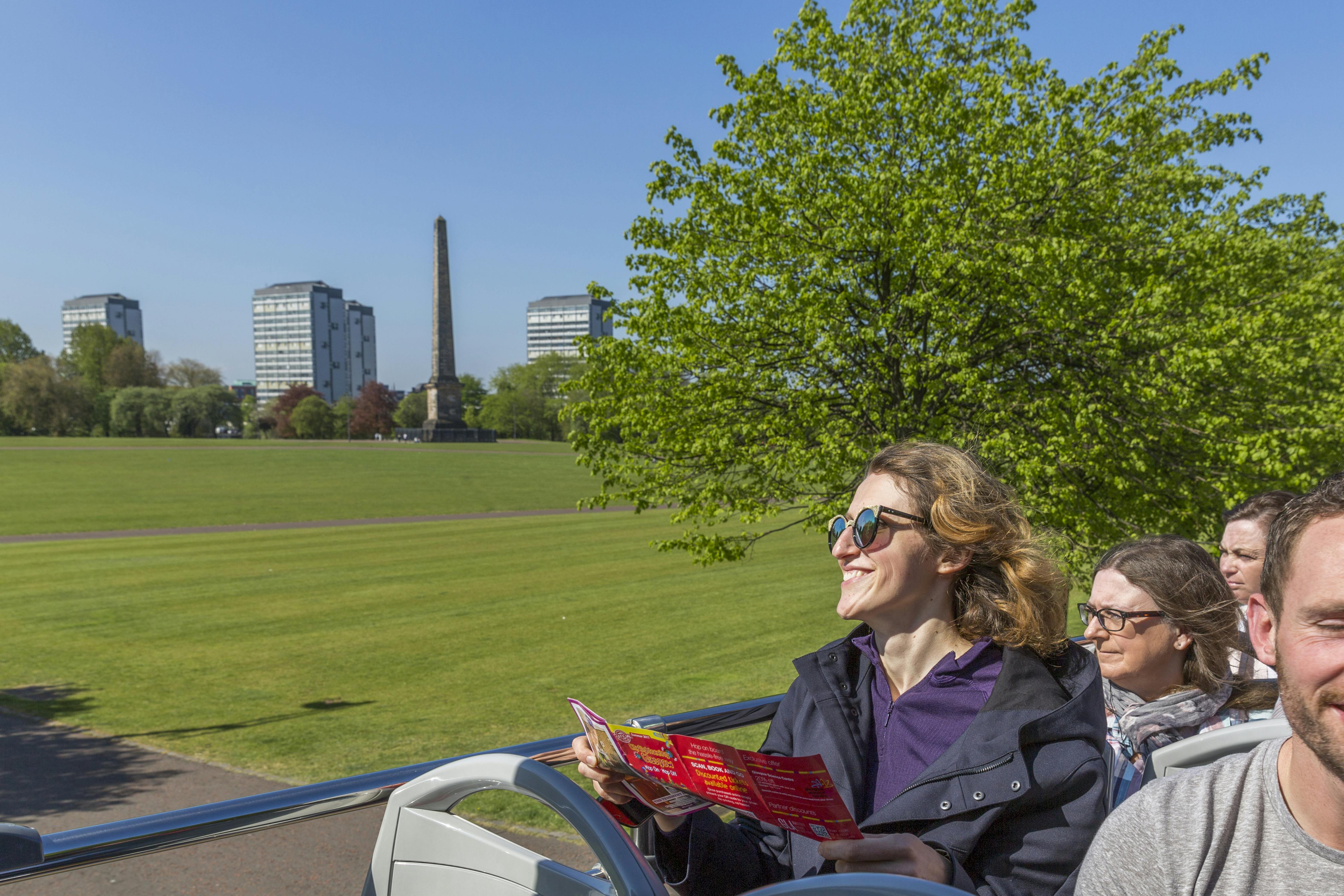 Άνθρωποι κάθονται σε ένα διώροφο λεωφορείο με ένα καταπράσινο πάρκο και δέντρα στο βάθος, ο ένας κρατώντας ένα φυλλάδιο και χαμογελώντας.