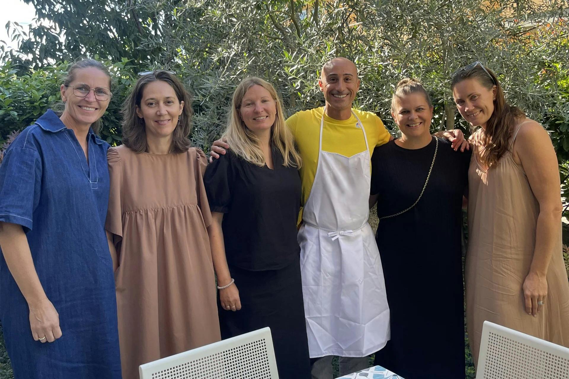 屋外に立って笑顔の5人のグループ。 黄色いシャツを着た人がエプロンを着ています。 木や屋外の椅子が見えます。