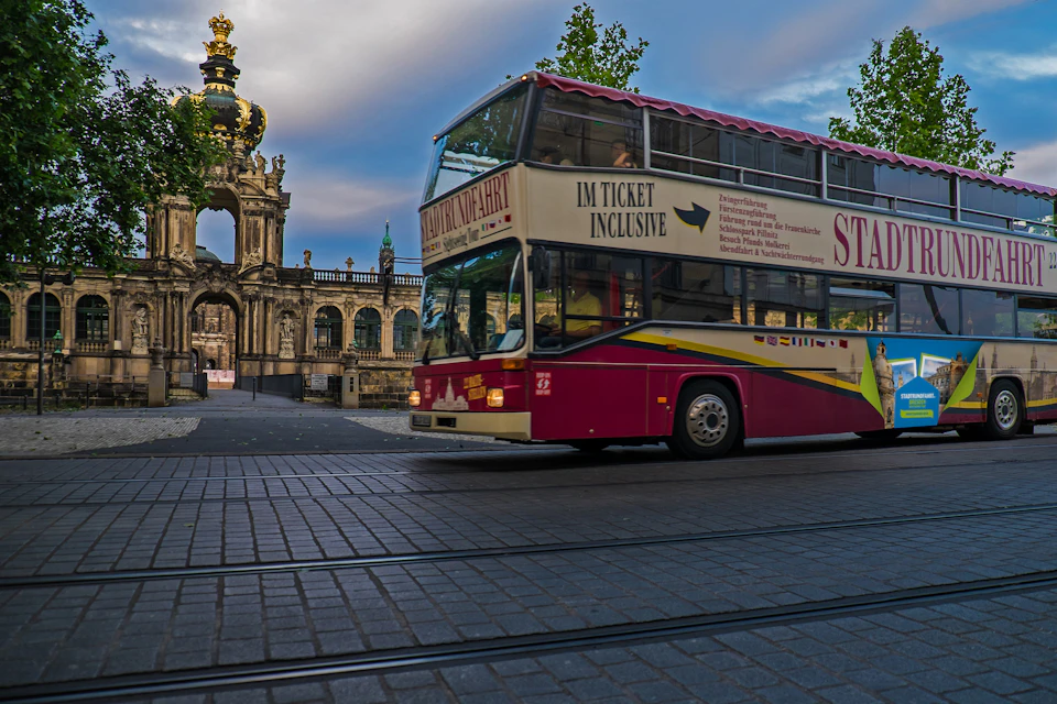 A red double-decker tour bus passes in front of a historic building with a crown-topped entrance under a partly cloudy sky.
